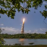 Ariane 6 rocket lifting off from Europe's Spaceport in French Guiana, seen through trees across a lake